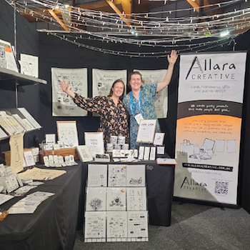 Two Allara Creative stallholders with arms raised in front of their market display of cards, prints and gifts.
